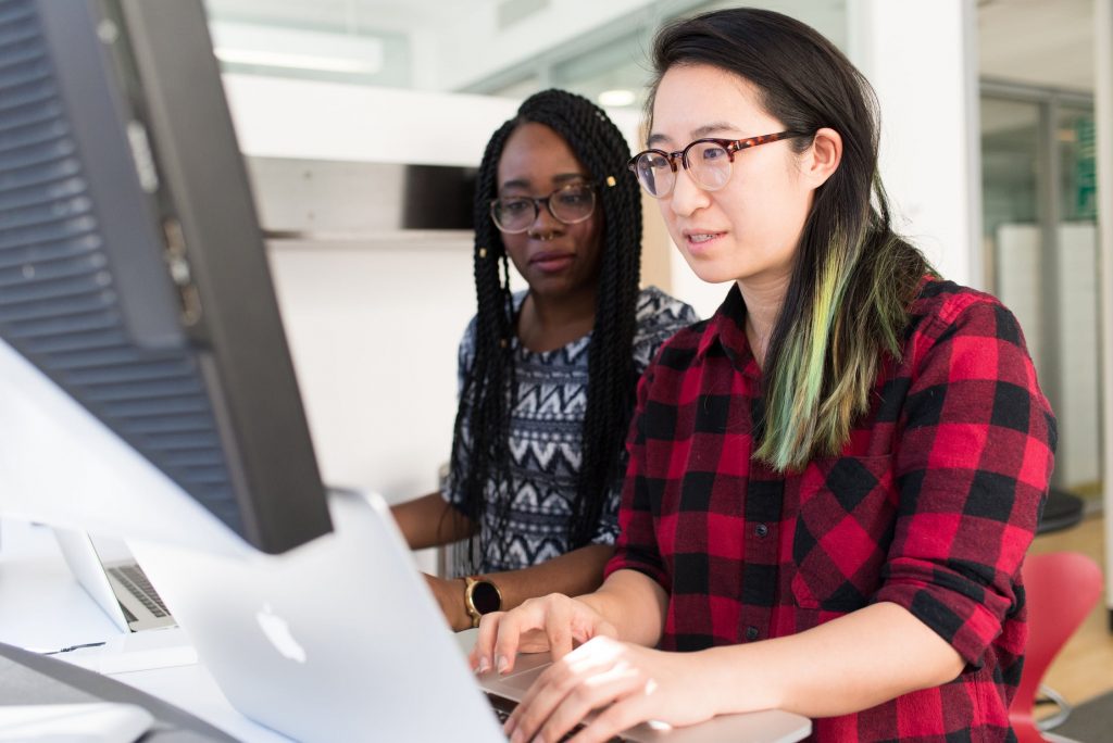 Asian and Black programming bootcamp students review technical assignment together, in front of computer screen.